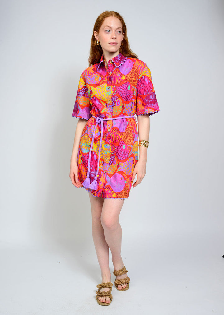 A woman with long red hair poses in the Zertz SCHOOL OF FISH DRESS, featuring vibrant ocean-inspired pink, orange, and purple abstract patterns and a belted waist. She pairs it with tan sandals, radiating coastal charm against a plain background.