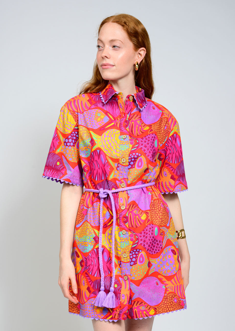 A woman with long red hair models the Zertz SCHOOL OF FISH DRESS, a vibrant short-sleeve design in pink, orange, yellow, and purple fish prints. The ocean-inspired look features a matching rope belt with tassels. She faces left against a plain background.