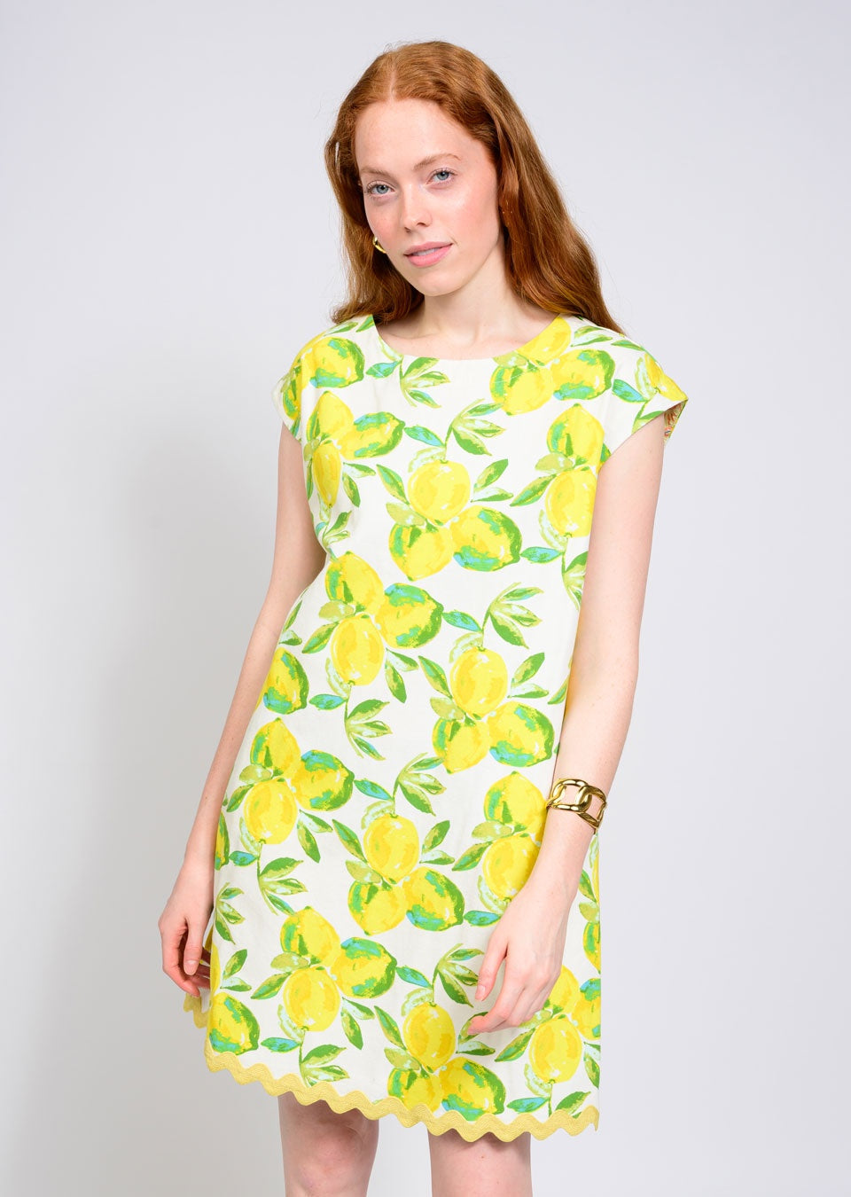 A woman with long red hair wears the Zertz LEMONADE DRESS, a white summer dress with bright yellow lemon print and green leaves, paired with a gold bracelet as she stands relaxed against a plain light background.