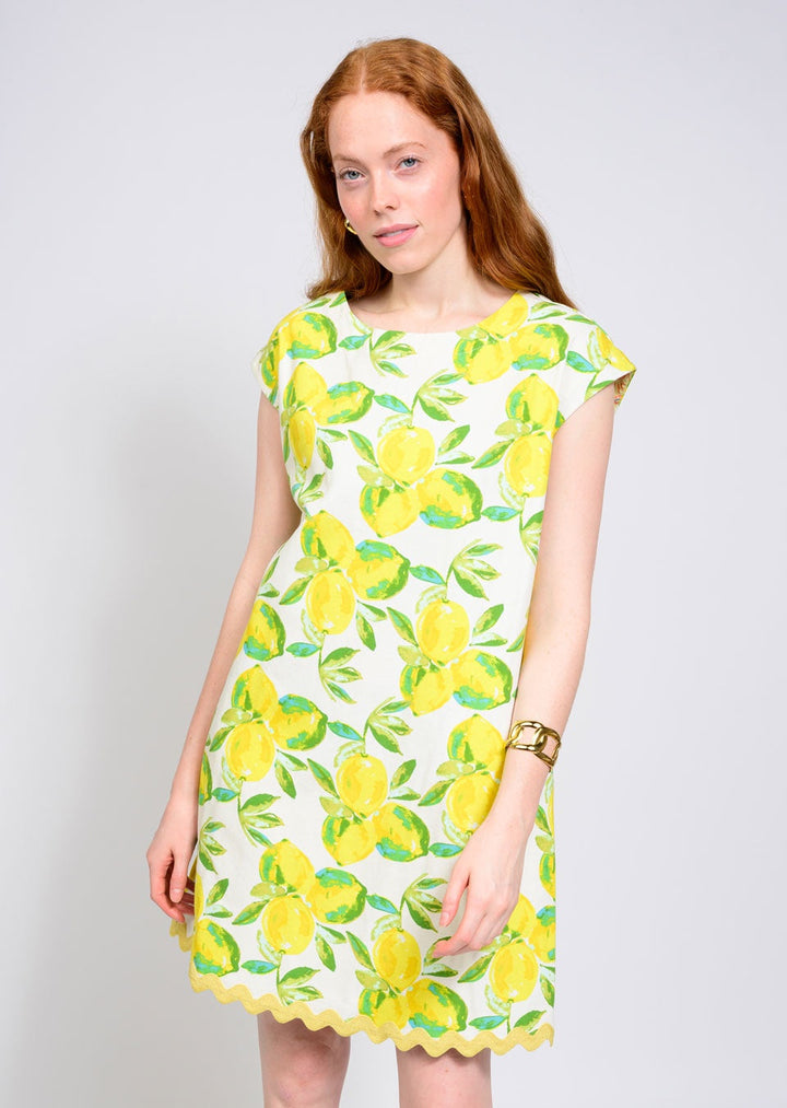 A woman with long red hair wears the Zertz LEMONADE DRESS, a white summer dress with bright yellow lemon print and green leaves, paired with a gold bracelet as she stands relaxed against a plain light background.