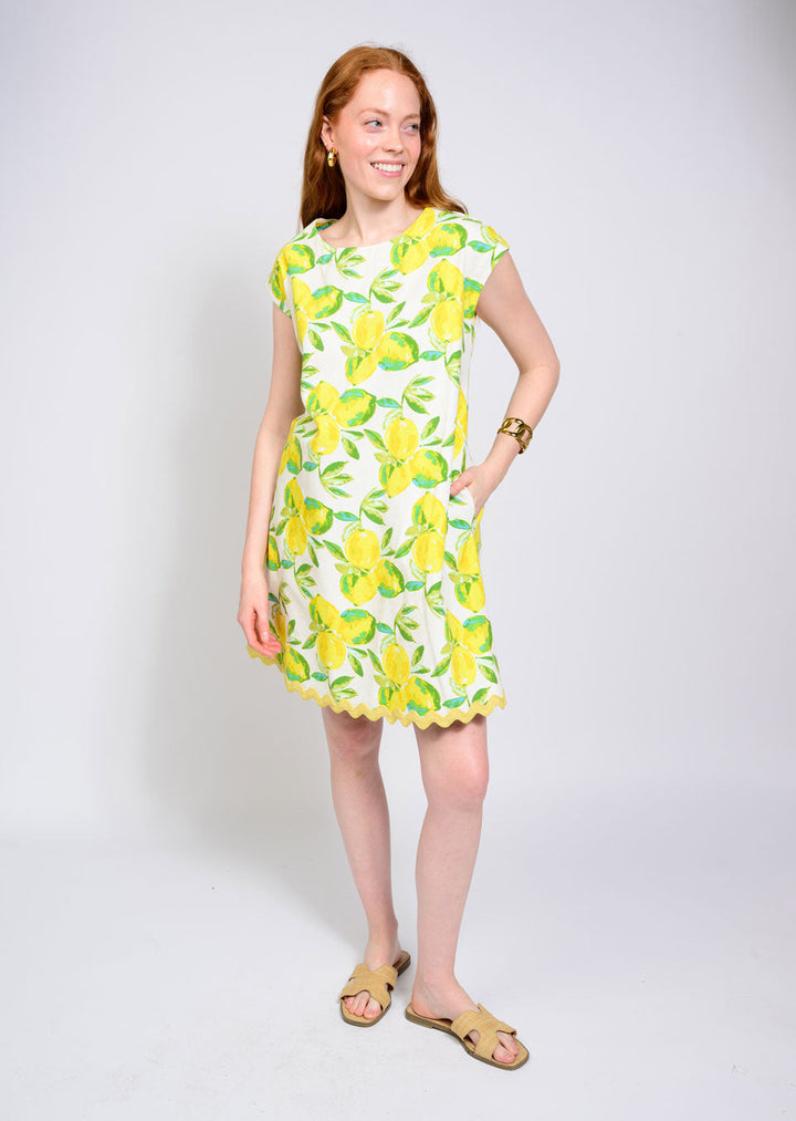 A woman with long red hair smiles as she poses against a plain light background, wearing the Zertz LEMONADE DRESS, featuring a vibrant yellow lemon print, paired with beige sandals and gold accessories.