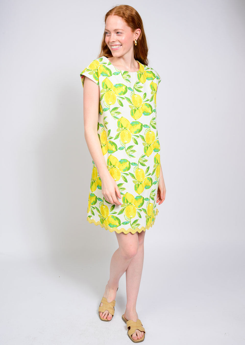 A woman with long red hair smiles in the Zertz LEMONADE DRESS—a short-sleeved, knee-length white dress printed with yellow lemons and green leaves. She stands on a white background and wears light-colored slide sandals.