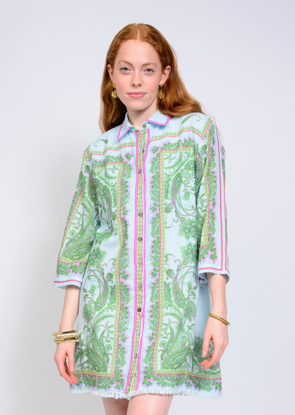 A woman with long red hair wears the Zertz PAISLEY SHIFT DRESS, a green paisley print button-up with pink borders and a relaxed silhouette, styled with gold hoop earrings and bracelets against a plain light background.