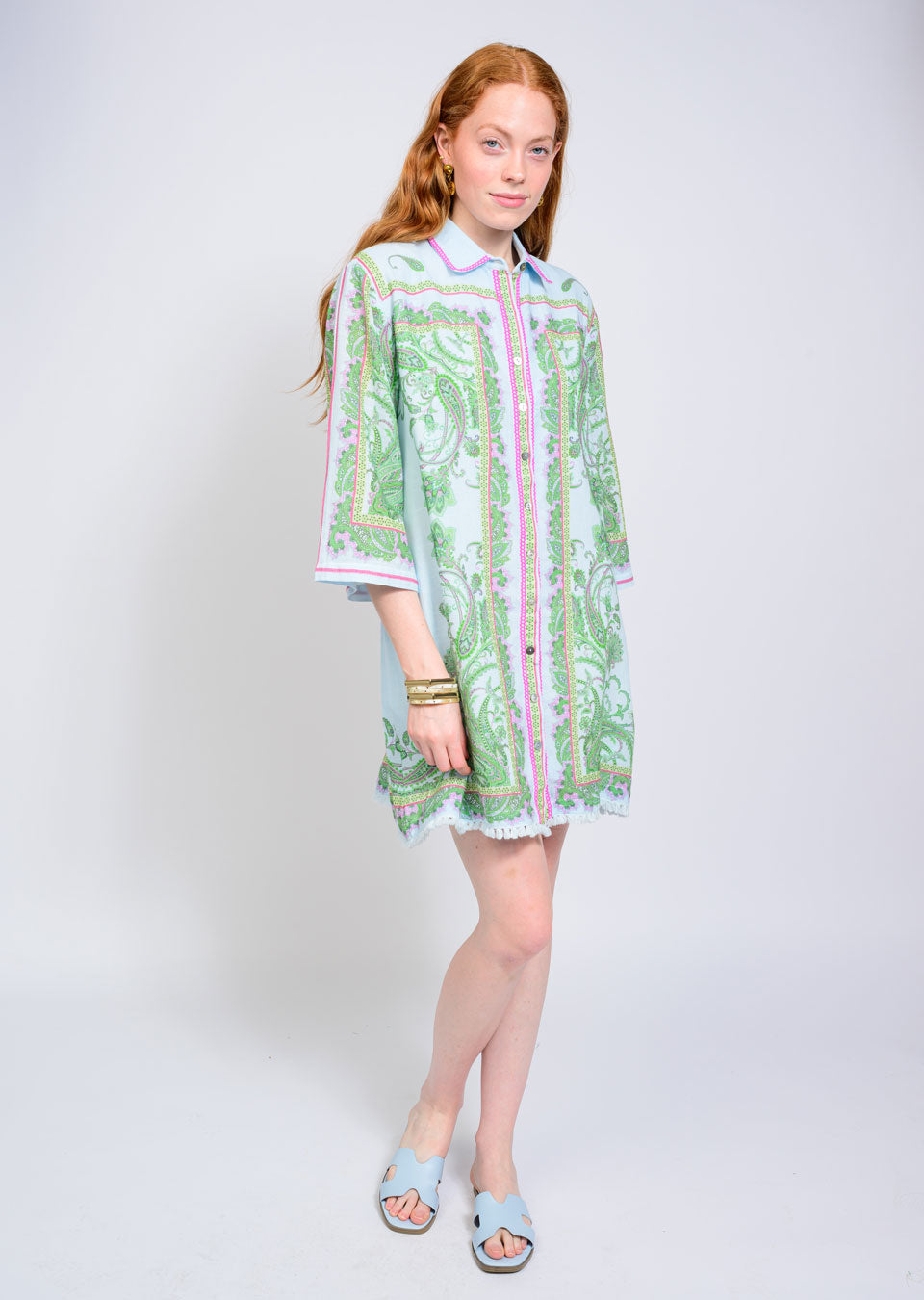 A woman with long red hair stands against a plain background, wearing the Zertz PAISLEY SHIFT DRESS—knee-length with a green and pink paisley print, white trim, and a relaxed silhouette. She pairs it with blue slide sandals and bracelets for versatile style.