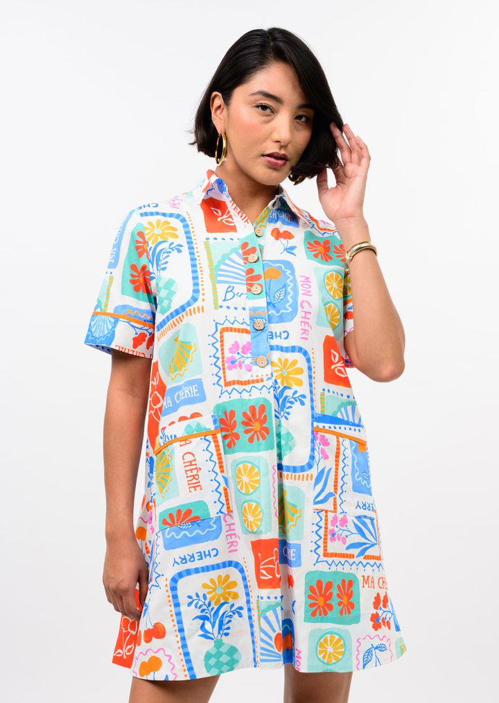 A woman with medium-length dark hair poses against a white backdrop in the Zertz FRUIT PATCH DRESS—a vibrant, short-sleeve button-down featuring bold floral and fruit patterns—touching her hair to highlight this playful statement piece.