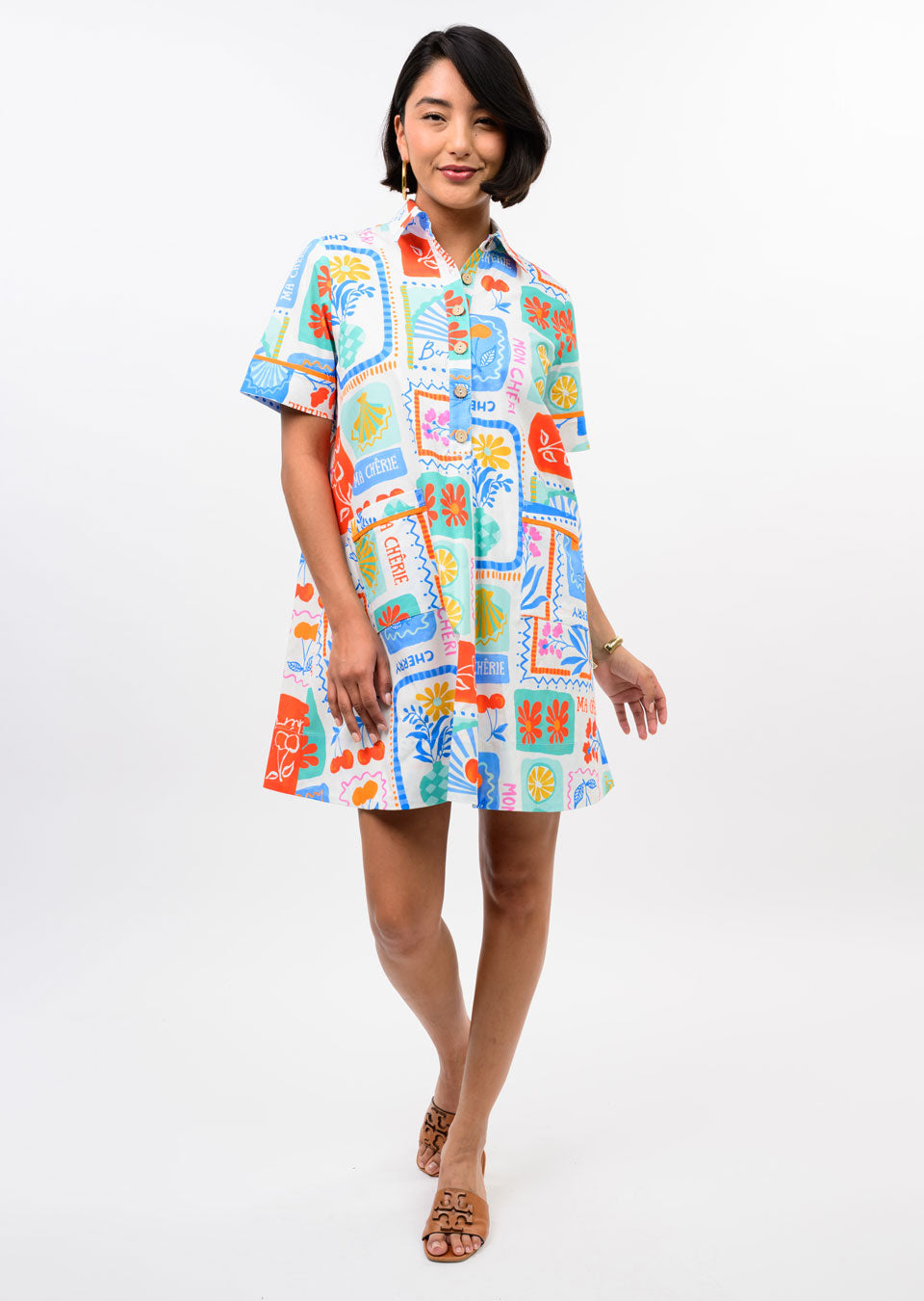 A woman models the Zertz FRUIT PATCH DRESS, a colorful short-sleeve button-up A-line dress with playful fruit prints. She stands smiling against a white background, one hand by her side and the other bent, wearing tan sandals.