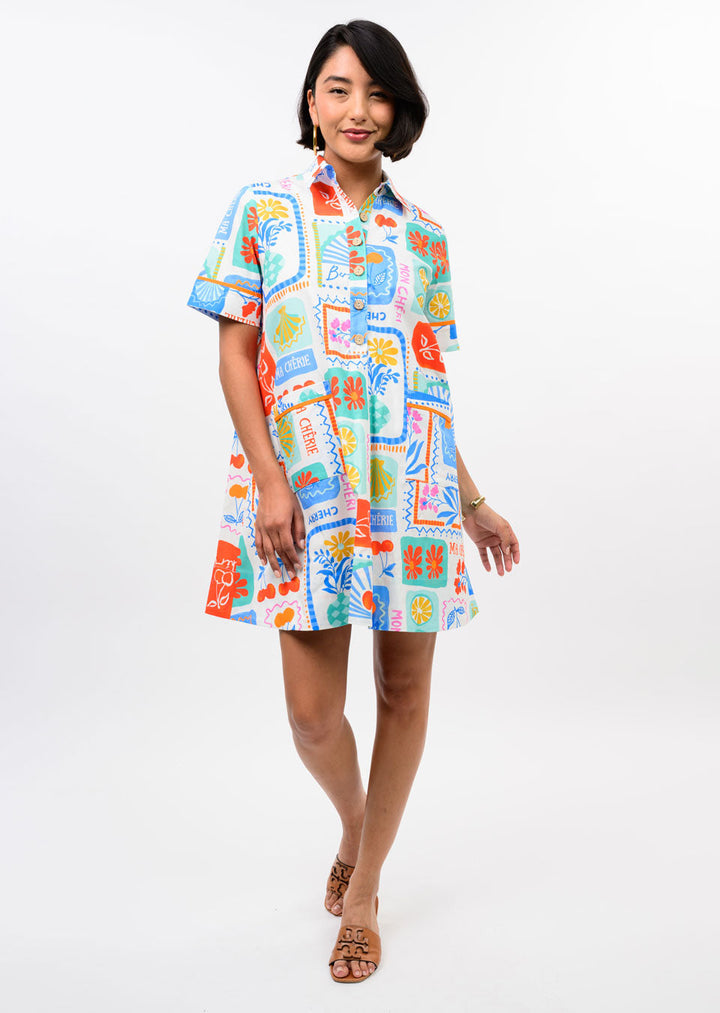 A woman models the Zertz FRUIT PATCH DRESS, a colorful short-sleeve button-up A-line dress with playful fruit prints. She stands smiling against a white background, one hand by her side and the other bent, wearing tan sandals.