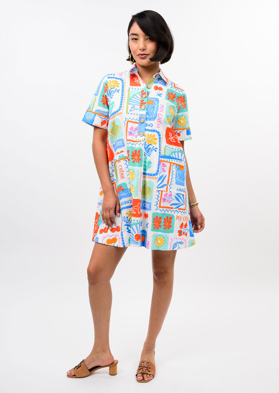 A woman models the Zertz FRUIT PATCH DRESS, featuring short sleeves and vivid fruit graphics. Paired with tan heeled sandals and simple jewelry, this standout piece shines against a plain white background.