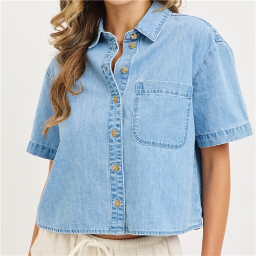 A woman wears The Jen Denim Top by Loca on Main, a light blue, short-sleeve, relaxed fit shirt with a chest pocket, paired with light-colored pants. Her face is not visible in the image.