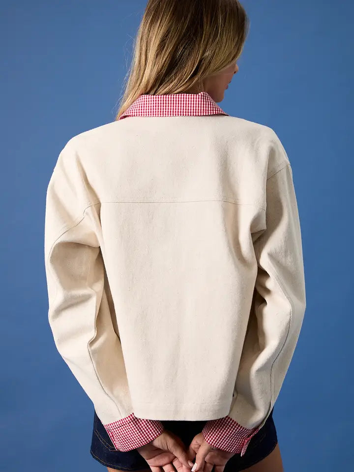 A woman stands against a blue background, facing away. She wears the Zertz USA FLAG BARN JACKET in cream with red and white checkered collar and cuffs, made from durable canvas, paired with dark shorts. Her hands are clasped behind her back.