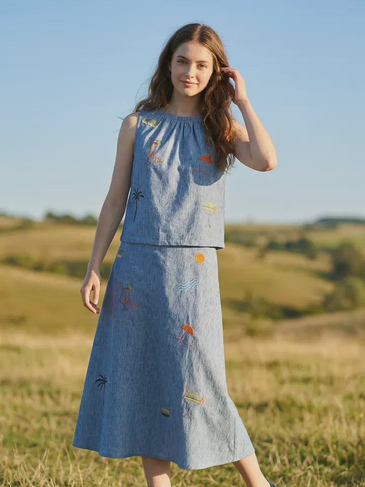 A young woman stands in a grassy field wearing the Zertz SUNSET EMBROIDERY SET, a sleeveless blue top and matching skirt with embroidered patches, set against rolling green hills beneath a clear blue sky.
