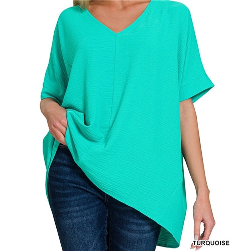 A woman wears the loose-fitting Airflow Basics Top by Loca on Main, featuring a turquoise color, V-neckline, lightweight fabric, asymmetrical hem, and oversized fit, paired with dark blue jeans for a relaxed look.