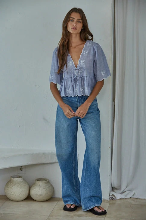 A woman with long brown hair wears the Zertz CELESTINE WOVEN TOP in light blue and wide-leg blue jeans. She stands indoors on a tiled floor near ceramic vases and a white curtain, looking at the camera.