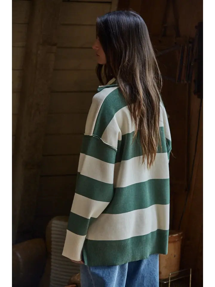 A person with long brown hair wears the Zertz FONTAINE HALF ZIP SWEATER, featuring green and white stripes, and blue jeans. They stand indoors facing away from the camera against a rustic wooden background.