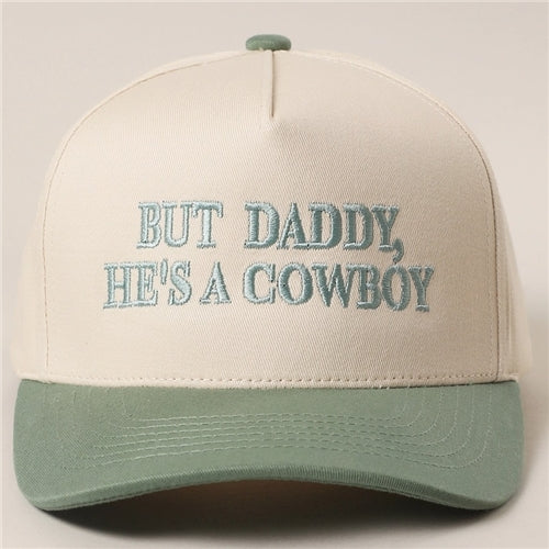 The Loca on Main "He's A Cowboy Trucker Hat" is a beige and light green two-tone hat with "BUT DADDY, HES A COWBOY" embroidered in light blue. One size fits most for comfortable wear.