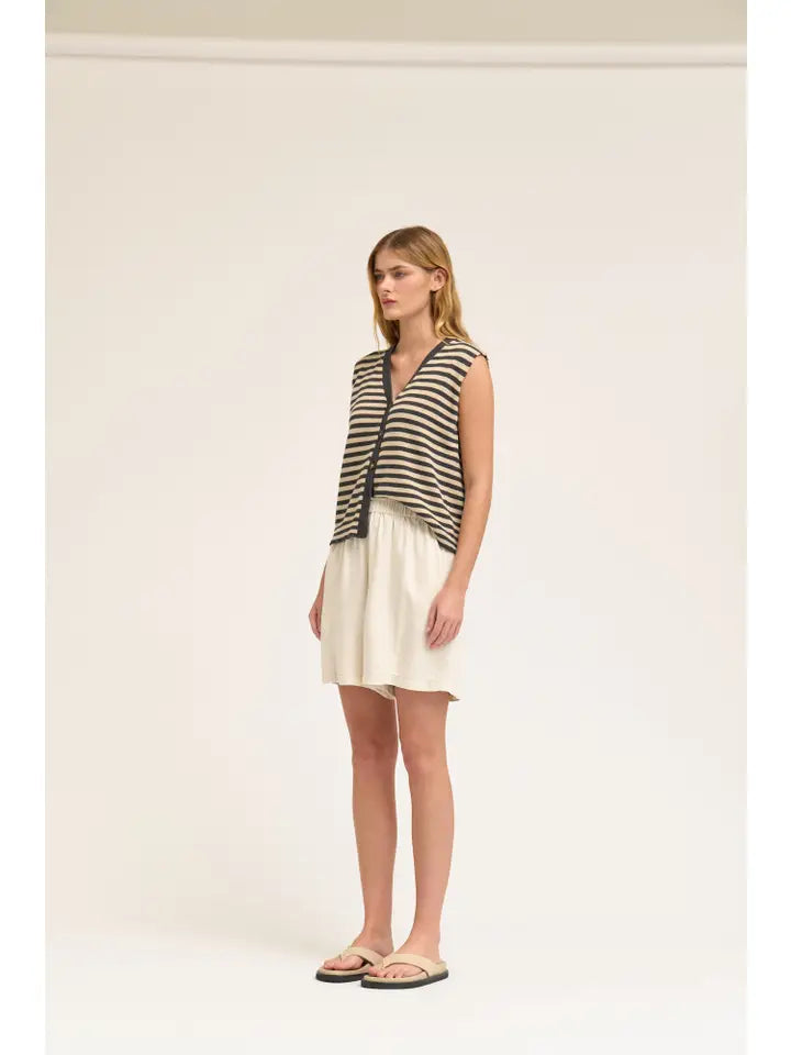A woman stands in a studio wearing Zertz TENCEL SHORTS in white, styled with a sleeveless black and beige striped knit vest and beige flip-flop sandals. The background is plain and light-colored.