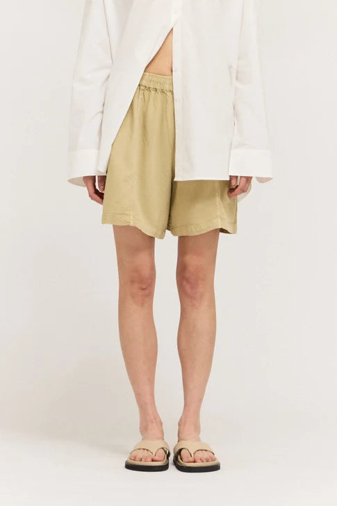 A person models Zertz TENCEL SHORTS—beige, breathable, and paired with a loose white shirt and beige sandals—standing against a plain white background. The image is cropped at the shoulders, showing only the torso, legs, and feet.