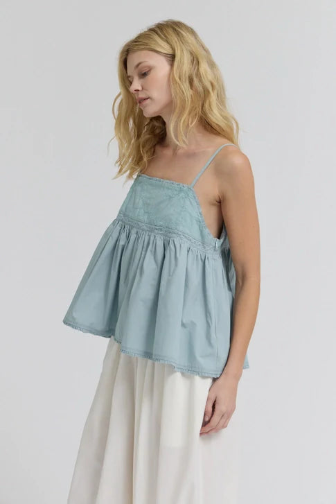 A woman with wavy blonde hair wears the Zertz EMBROIDERED SLEEVELESS TOP, a loose light blue piece in breathable fabric, paired with white flowy pants. She stands against a plain light background, looking down with a relaxed expression.