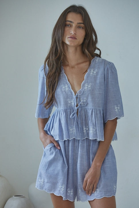 A woman with long brown hair wears the Zertz CELESTINE WOVEN TOP in light blue with white floral embroidery—an airy, effortless staple—paired with matching shorts, standing before a plain, light-colored background.