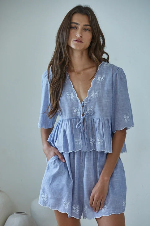 A woman with long brown hair models the CELESTINE SHORTS by Zertz—a light blue, short-sleeve romper with scalloped edges and white floral embroidery—standing relaxed against a light wall.