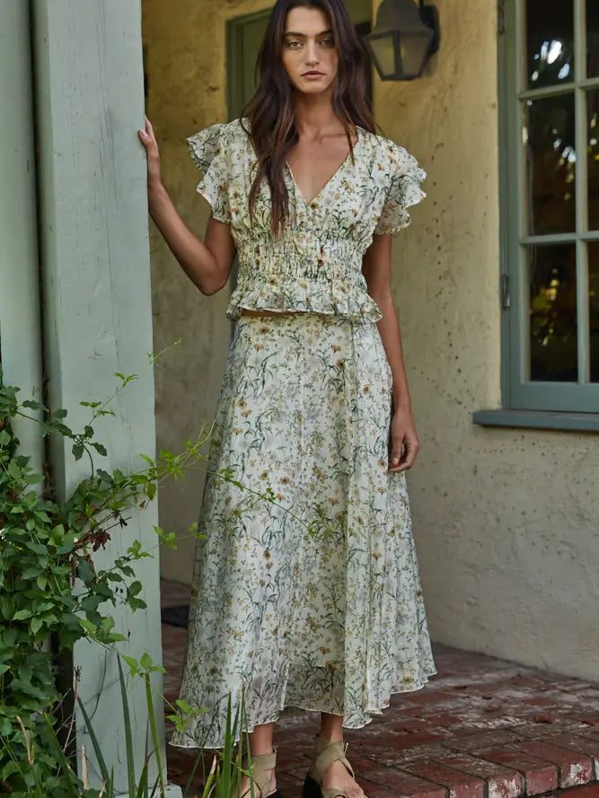 A woman with long brown hair and a neutral expression stands on a brick porch by a green pillar, wearing the Zertz SEREIA FLORAL TOP with a matching midi skirt. Green plants nearby enhance the fresh, natural vibe.