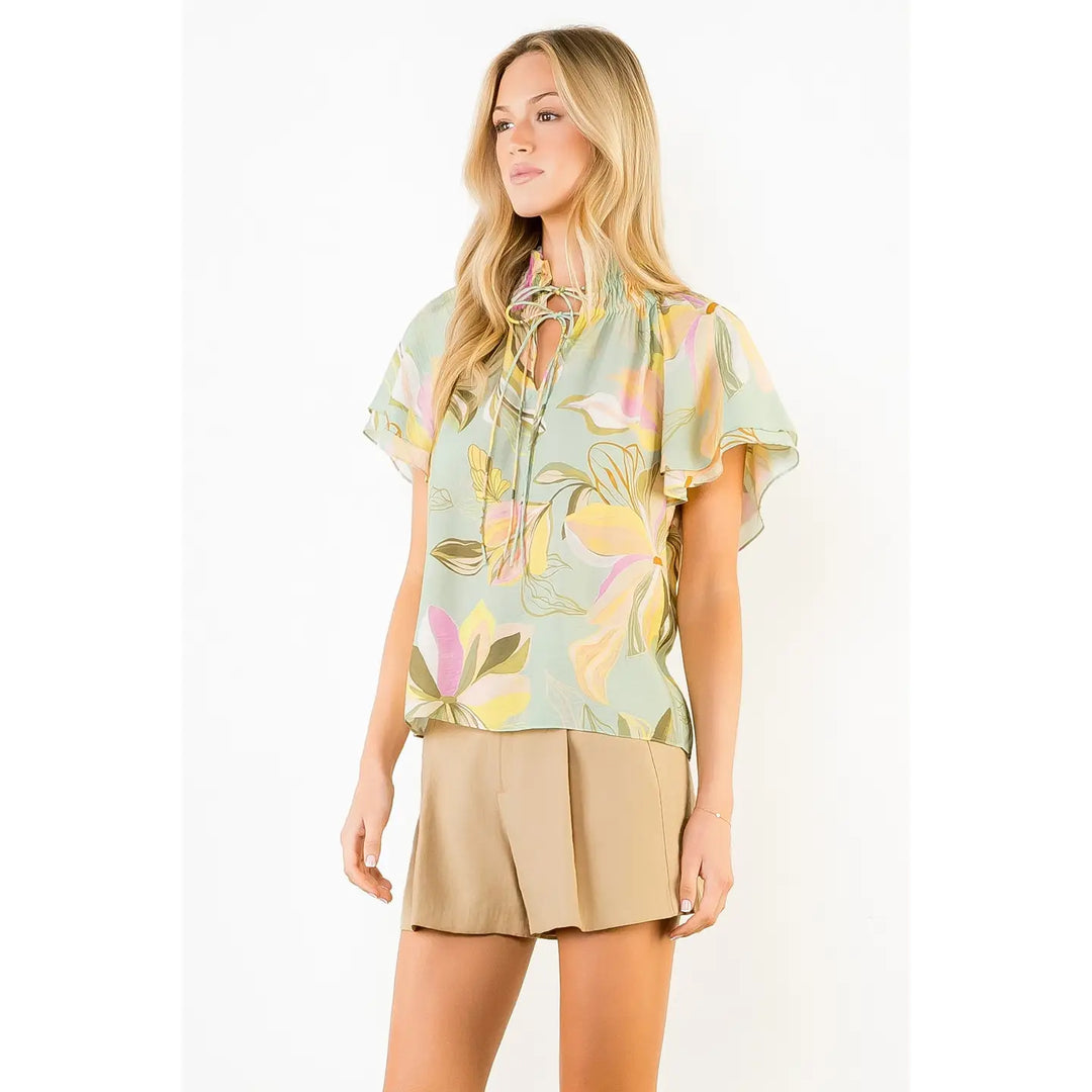 A woman wears the Zertz FLOWY FLOWER TOP WITH TIE FRONT in a pastel floral design, paired with tan pleated shorts, standing against a plain white background.