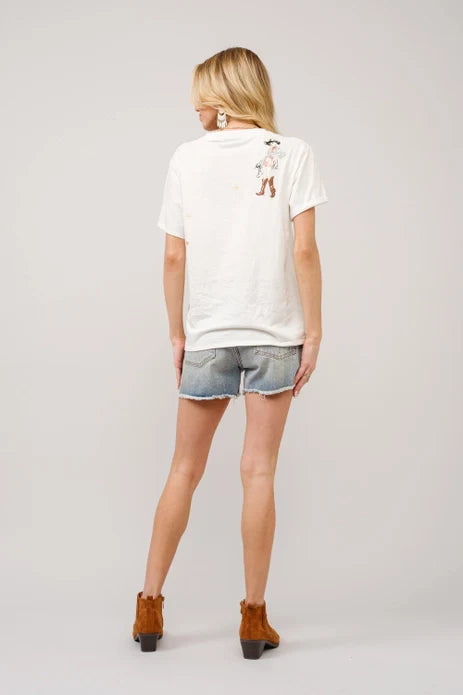A woman faces away in a Zertz WESTERN GRAPHIC T SHIRT featuring a small cowboy graphic on the back, paired with denim cut-off shorts and brown ankle boots, posed against a plain light background.