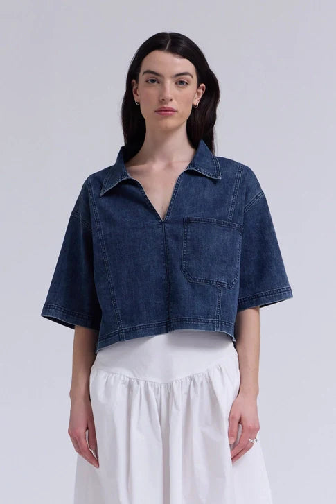 A woman with long dark hair models the Zertz DENIM CROP SHIRT, featuring a chest pocket, paired with a white gathered skirt against a plain light background.