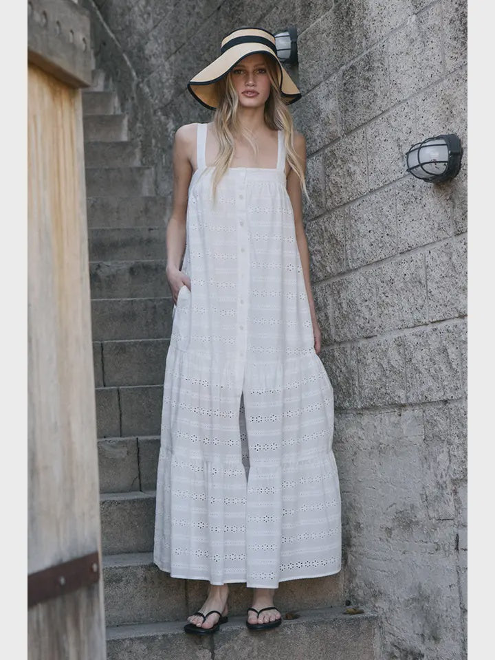 A woman wears the Zertz EYELET BUTTON FRONT MAXI DRESS, a sleeveless white summer dress, with a wide-brimmed hat and black flip-flops, standing on stone steps by a concrete wall with outdoor light fixtures.