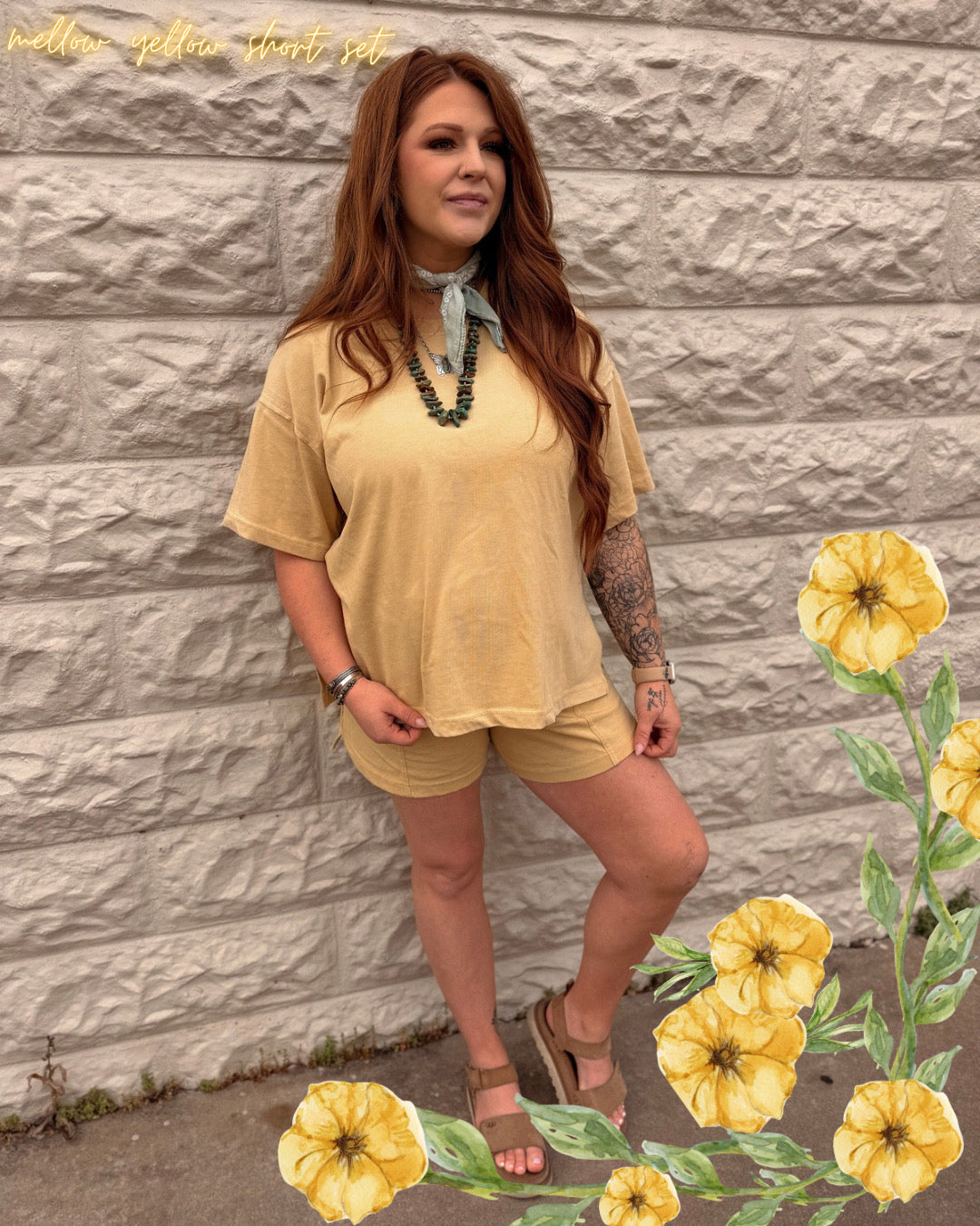 A woman with long brown hair models The Mellow Yellow Set by Loca on Main—an everyday lounge look with relaxed-fit shorts. She pairs it with sandals, a scarf, and jewelry, standing by a stone wall with illustrated yellow flowers in the corner.
