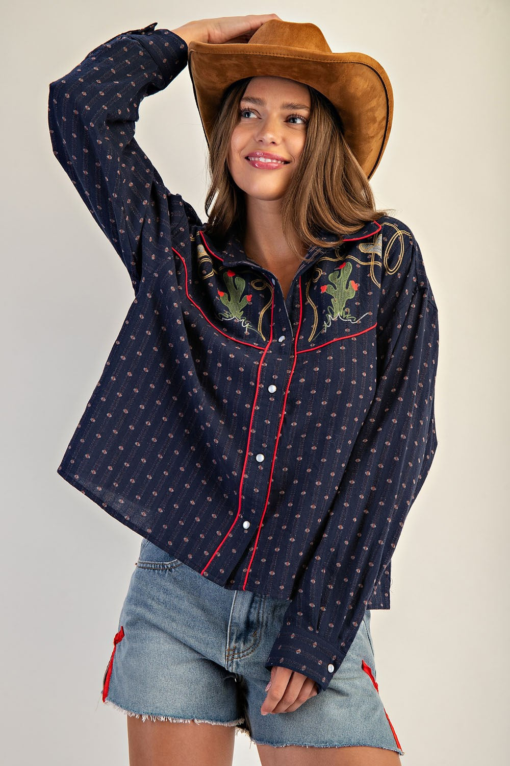 A smiling woman in a brown cowboy hat wears the Zertz WESTERN BUTTON DOWN SHIRT, featuring navy blue fabric with Western embroidery, paired with light denim shorts as she poses with one arm raised against a neutral background.