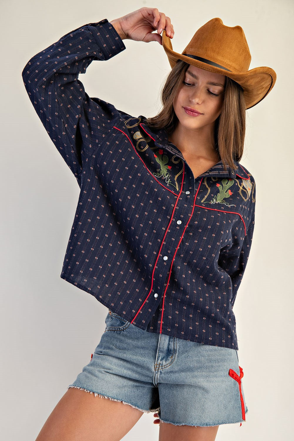 A woman wearing Zertz WESTERN BUTTON DOWN SHIRTS in navy with floral embroidery, a brown cowboy hat, and denim shorts with red ribbon detail, poses holding her hat and looking down.