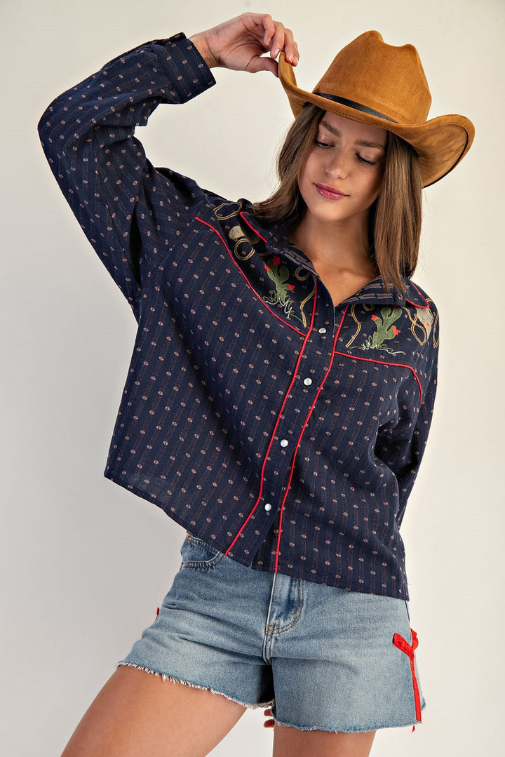 A woman wearing Zertz WESTERN BUTTON DOWN SHIRTS in navy with floral embroidery, a brown cowboy hat, and denim shorts with red ribbon detail, poses holding her hat and looking down.