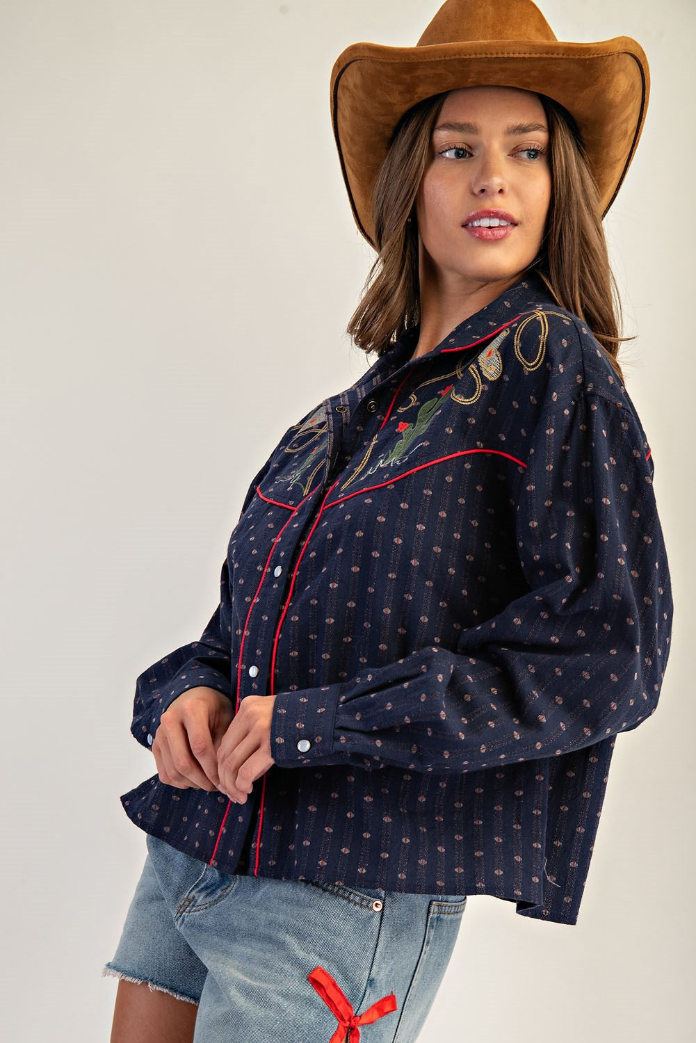 A woman in a brown cowboy hat, Zertz WESTERN BUTTON DOWN SHIRTS in navy blue with embroidery, and denim shorts featuring a red ribbon poses against a neutral background.