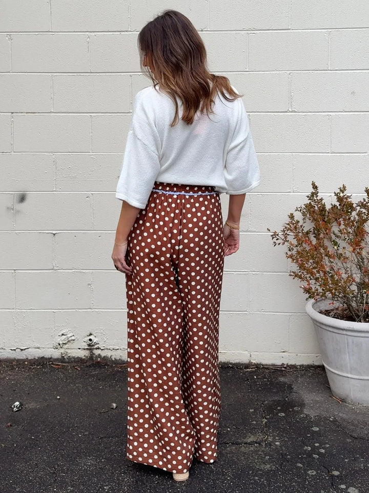 A woman stands with her back to the camera, wearing a white top and Zertz POLKA DOT PANTS—high-waisted, wide-leg brown pants with white polka dots—outdoors near a white brick wall and a potted plant.
