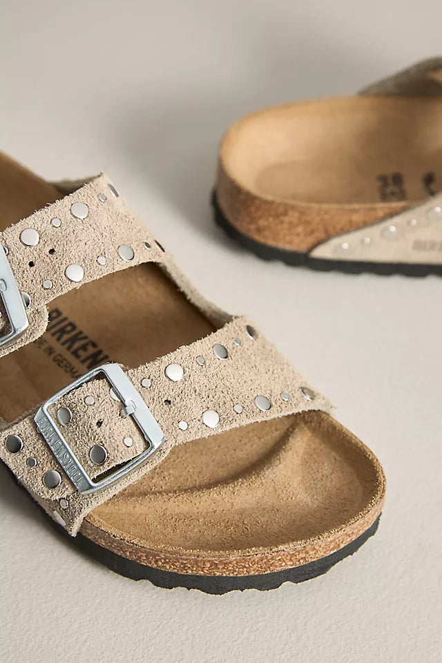 A close-up of Zertz ARIZONA RIVET SUEDE TAUPE N sandals in beige suede with two silver buckles, metallic studs on the straps, a cork footbed, and black rubber soles. Another sandal is partially visible in the background.