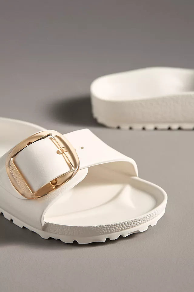 A close-up of the Zertz MADRID BB EVA EGGSHELL sandal, a white ultra-lightweight style with a thick strap and large gold buckle, displayed on a gray surface with its matching pair in the background.
