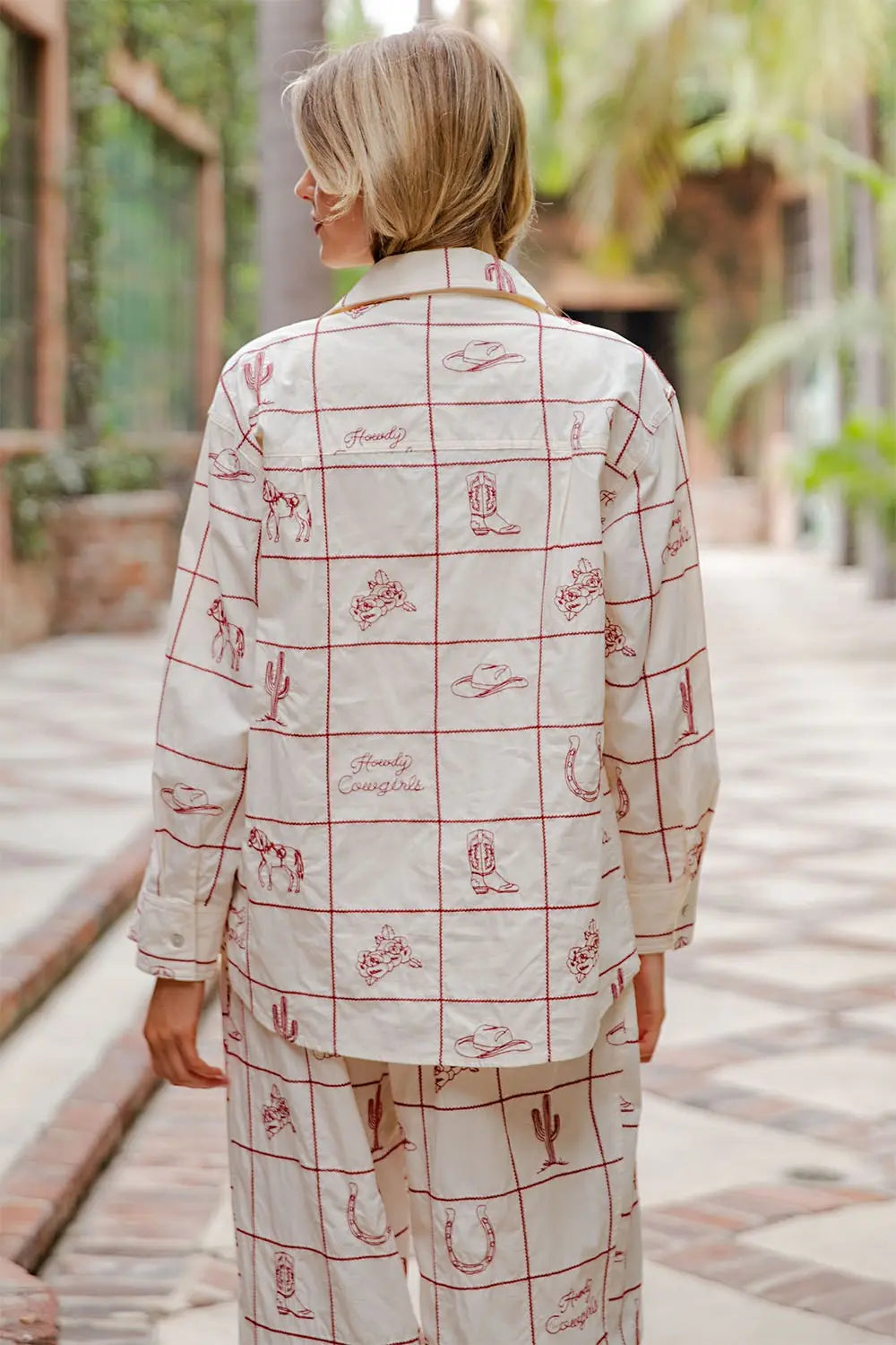 A person with short blonde hair wears the Zertz WESTERN EMB BUTTON DOWN and matching pants, featuring red desert-themed prints and grid lines, while standing outdoors on a tiled path surrounded by greenery and palm trees.