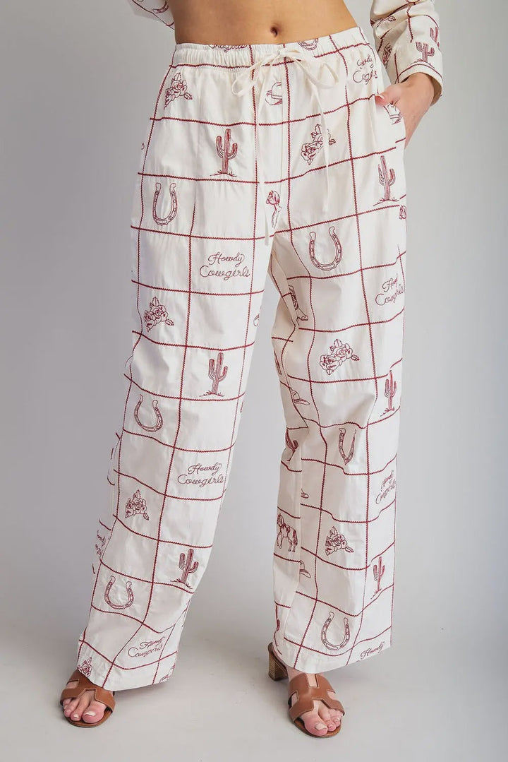 Zertz WESTERN EMB PANTS feature a white wide-leg design with a red grid, cacti, horseshoes, cowboy hats, boots, and “Howdy Cowgirl” embroidery. Styled with heeled sandals for a classic ranch look.