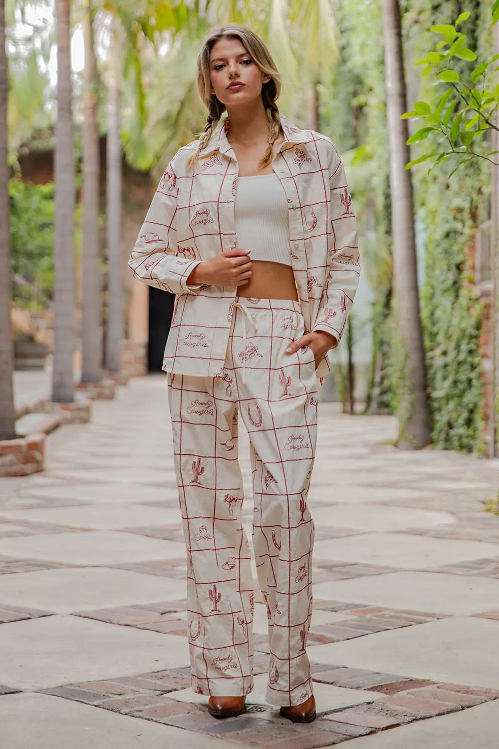 A woman stands outdoors in Zertz’s WESTERN EMB PANTS, a cream-colored ranch-style pantsuit with a grid pattern and red sketches, paired with a white crop top and brown shoes.