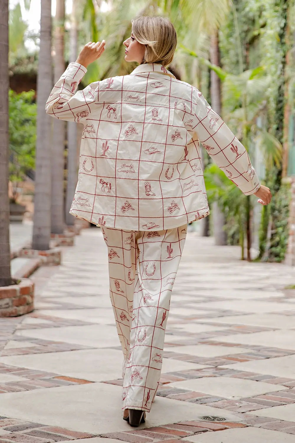 A person with blonde hair walks on a tiled path lined with palm trees, seen from the back, wearing Zertz’s WESTERN EMB PANTS—a cream suit featuring a red grid and desert-inspired designs.