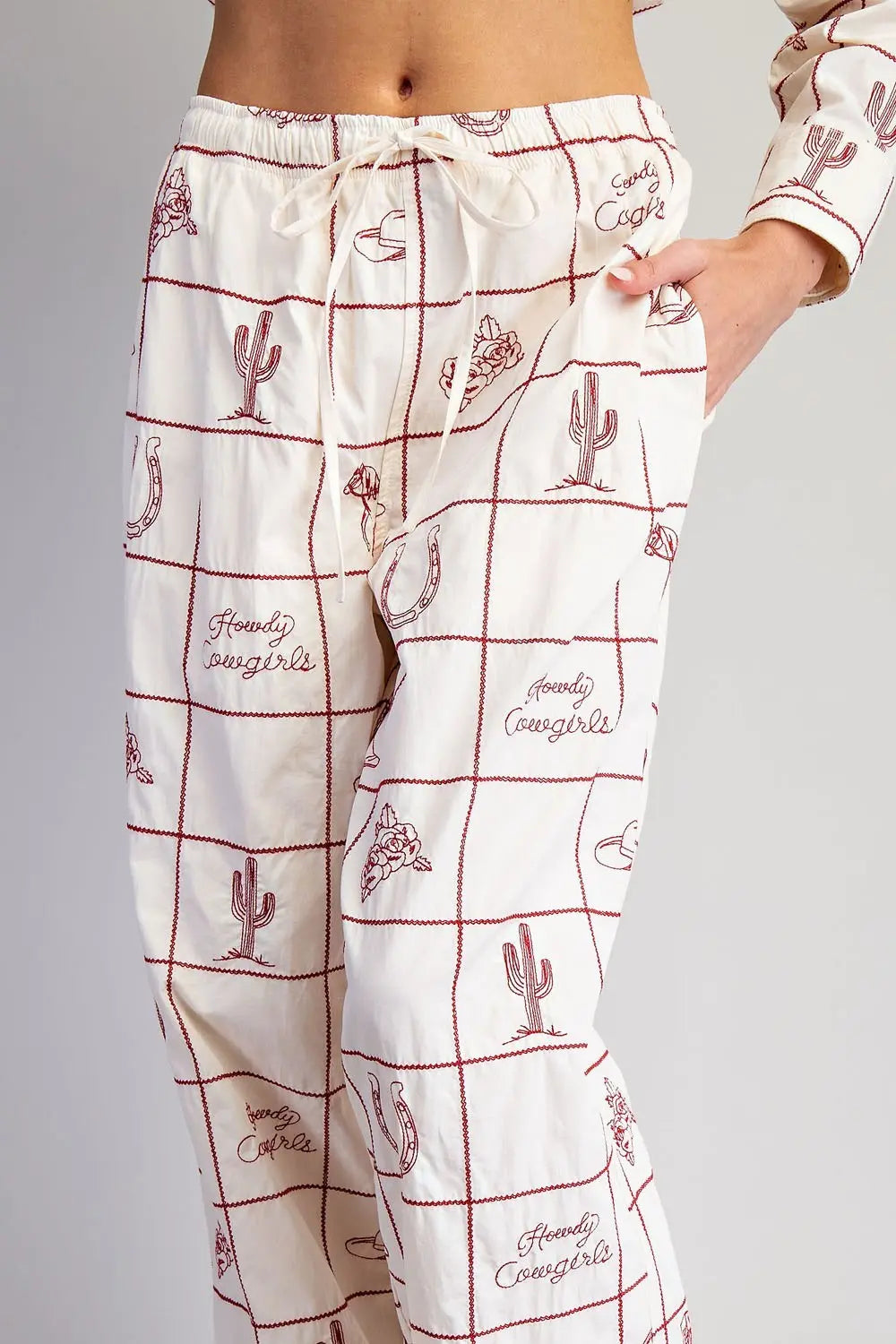 A person wears Zertz WESTERN EMB PANTS in cream, featuring red Western motifs—cacti, cowboy hats, horseshoes, boots, and “Howdy Cowgirl” text. One hand rests casually in the pocket for a laid-back ranch-inspired look.