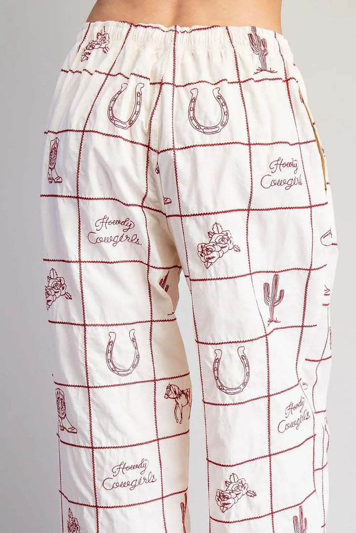 A person wears Zertz WESTERN EMB PANTS—white western pants with a red grid, ranch-themed embroidery like horseshoes, cacti, cowboy hats, and the phrase "Howdy Cowgirls.