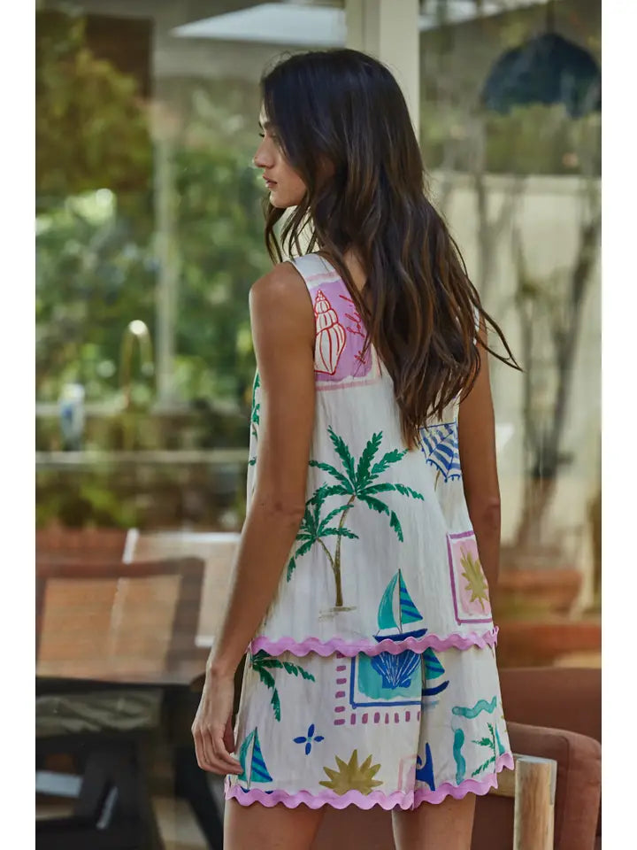 A woman with long brown hair stands outdoors in a sleeveless white dress with colorful palm trees, seashells, and sailboats, paired with Zertz MONTMARTRE WOVEN SHORTS for a relaxed and stylish summer look.
