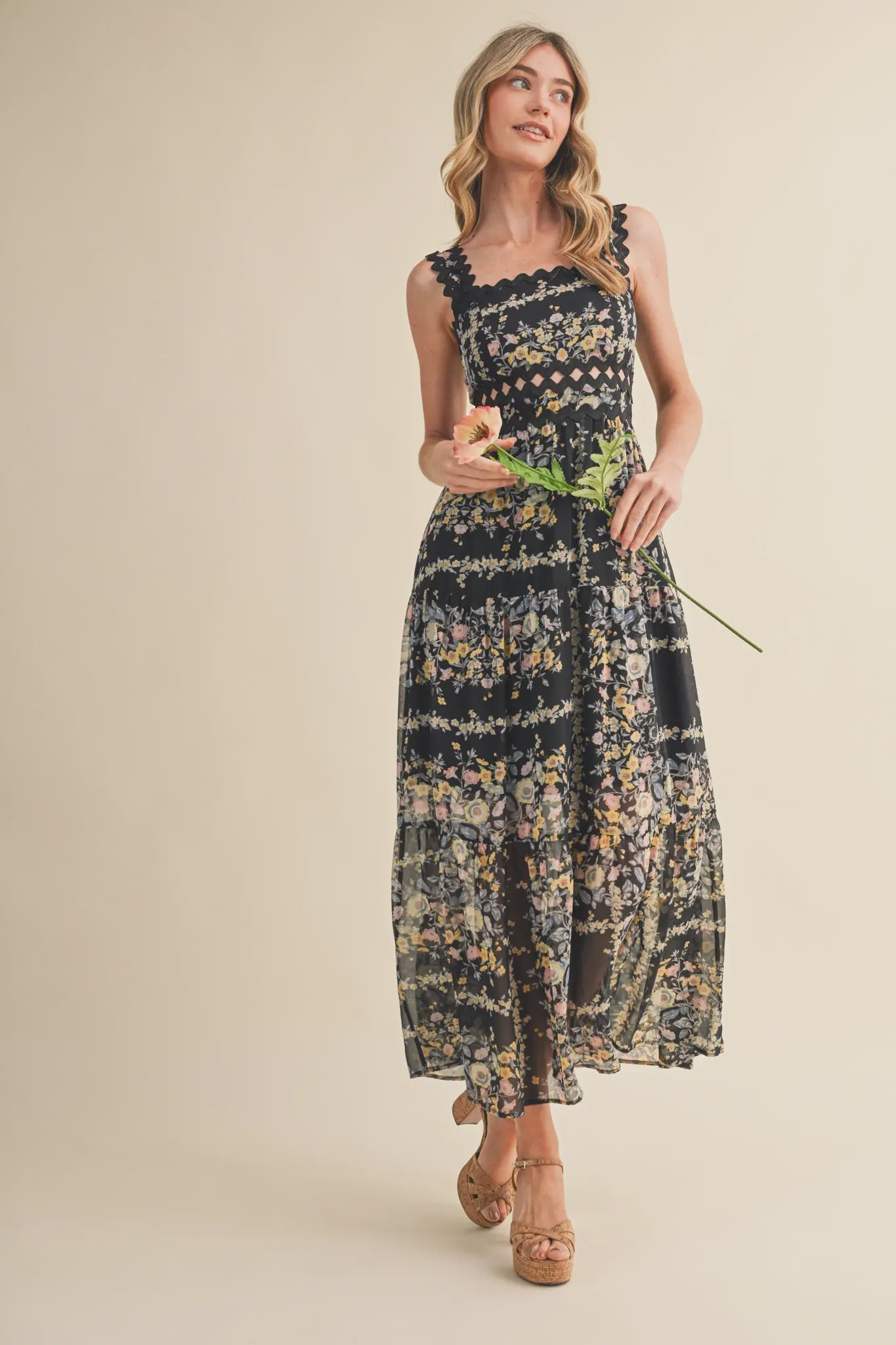 A woman in Zertz's PRINTED DRESS—sleeveless, ankle-length, navy with a flowing silhouette—holds a peach flower and smiles against a light beige background.