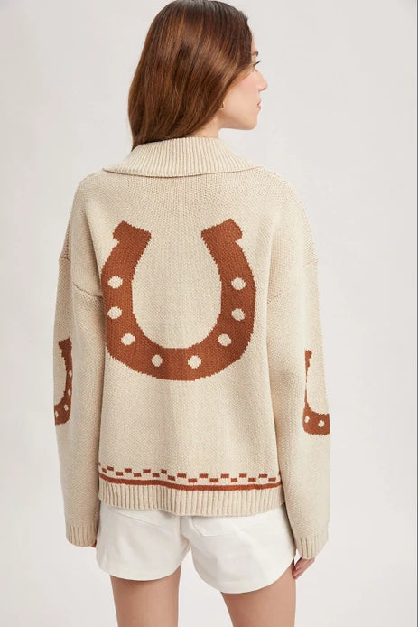 A woman with long brown hair models the Zertz HORSE GRAPHIC BUTTON CARDIGEN, which features bold horseshoe patterns on the back and sleeves. She pairs it with white shorts and stands against a simple light background.