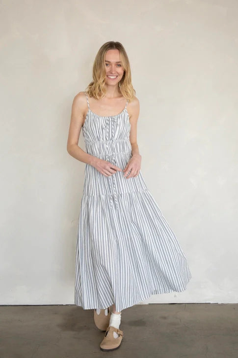 A woman with blonde hair smiles against a light wall, wearing the Zertz MAXI COTTON DRESS—a sleeveless white and blue striped outfit—paired with beige shoes and white socks.