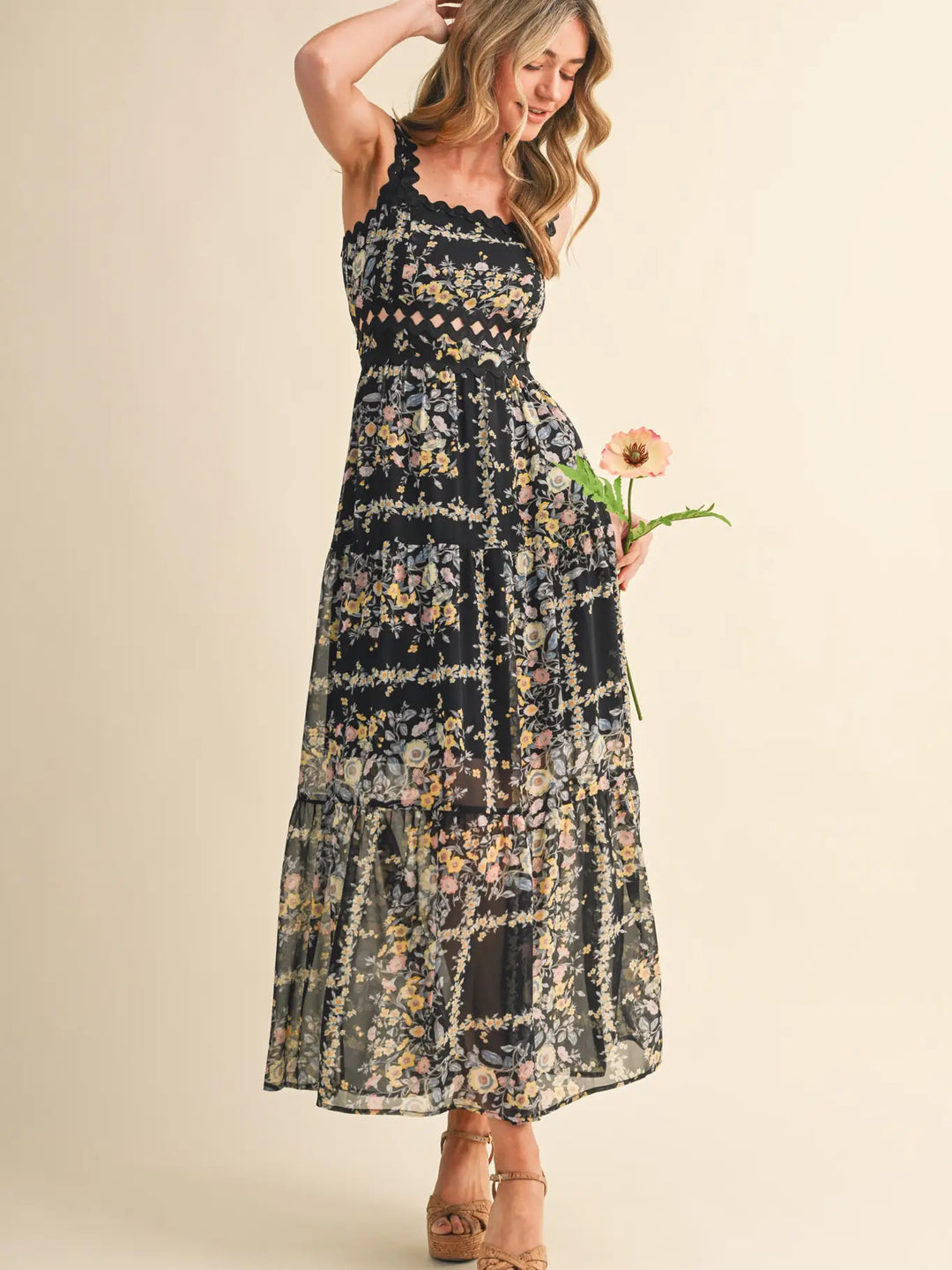 A woman in the Zertz PRINTED DRESS, featuring a sleeveless black print and flowing silhouette, stands on platform heels holding a pink flower and smiling gently against a neutral background.