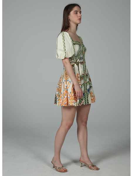 A woman stands in profile against a plain gray background wearing the Zertz CORA PUFF MINI DRESS, featuring green, blue, and orange tropical prints, puff sleeves, a square neckline, and paired with heeled sandals.