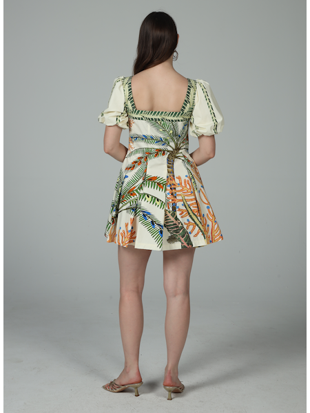 A woman stands with her back to the camera, wearing the Zertz CORA PUFF MINI DRESS in cream with a colorful tropical leaf and plant pattern, featuring puffed sleeves and paired with open-toed heels.